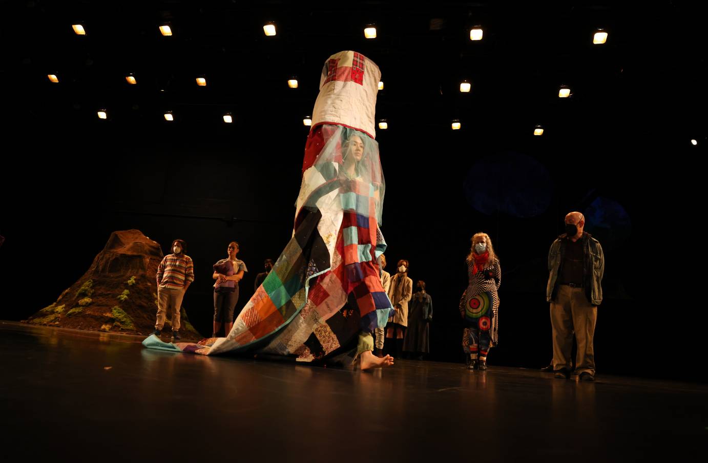 amongst a crowd of onlookers on stage walks a woman dressed in an elaborate quilt sculpture
